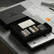 The full unboxing reveal of the OTTORON Midnight Industrial Master Sample Kit, showcasing the open matte black rigid box with double-layer black EVA foam, revealing premium cards, the Foil Specifier, the Adhesive Archive swatch book, and the $20 debossed credit card resting on top.